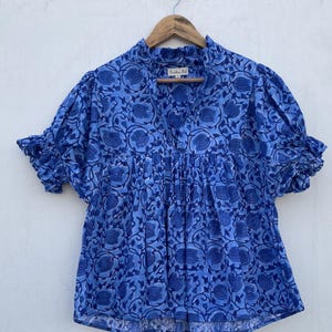 Puede incluir: Una blusa de algodón con estampado floral azul y blanco, cuello con volantes y mangas cortas abullonadas. La blusa tiene un corpiño fruncido y un ajuste holgado.
