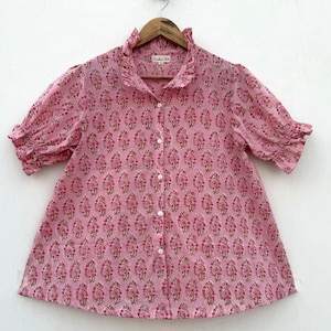 May include: A pink button-down shirt with a floral pattern. The shirt has short, ruffled sleeves and a ruffled collar. The floral design features pink and green accents. The shirt is hanging on a wooden hanger.