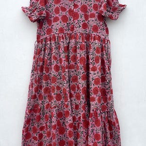May include: A pink maxi dress with a red fruit pattern. The dress has short, frilled sleeves, a button-down bodice, and a tiered skirt. The fabric features a light pink background with a repeating pattern of red fruit and green leaves.