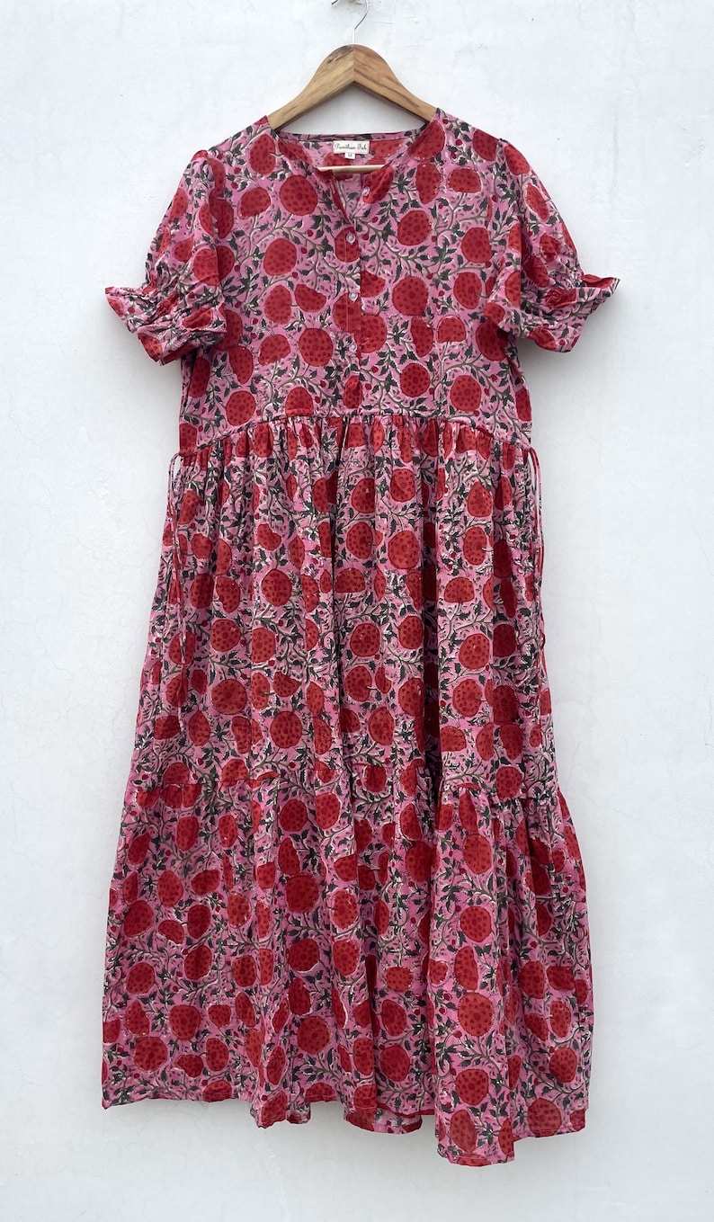 May include: A pink maxi dress with a red fruit pattern. The dress has short, frilled sleeves, a button-down bodice, and a tiered skirt. The fabric features a light pink background with a repeating pattern of red fruit and green leaves.