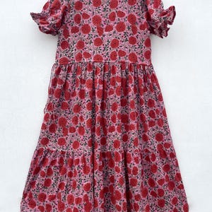 May include: A pink dress with a red floral print. The dress features short sleeves with frilled edges and a tiered skirt. The fabric is lightweight, possibly cotton. The dress is hanging on a wooden hanger.