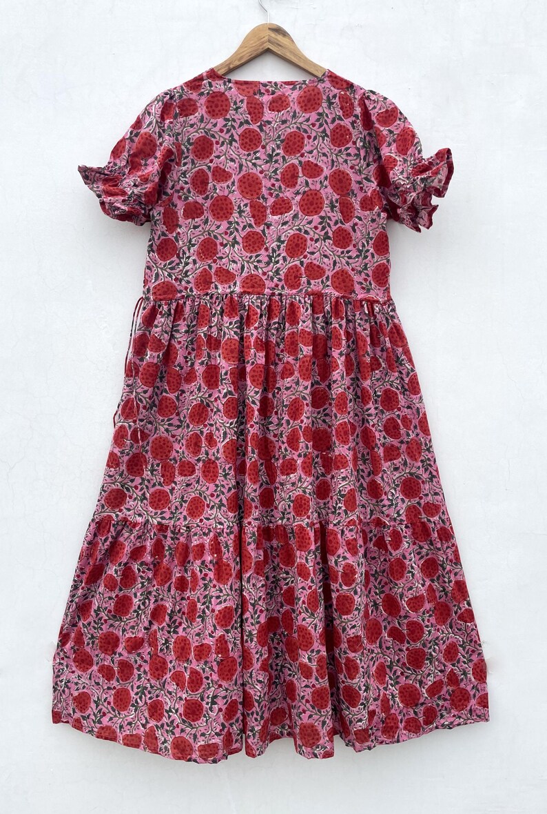 May include: A pink dress with a red floral print. The dress features short sleeves with frilled edges and a tiered skirt. The fabric is lightweight, possibly cotton. The dress is hanging on a wooden hanger.