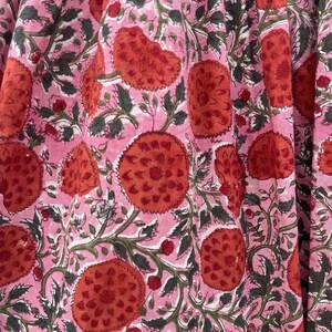 May include: A close-up of a pink fabric featuring a repeating pattern of large, stylized red flowers. The flowers have a textured appearance and are surrounded by green leaves and vines. The background is a soft pink.