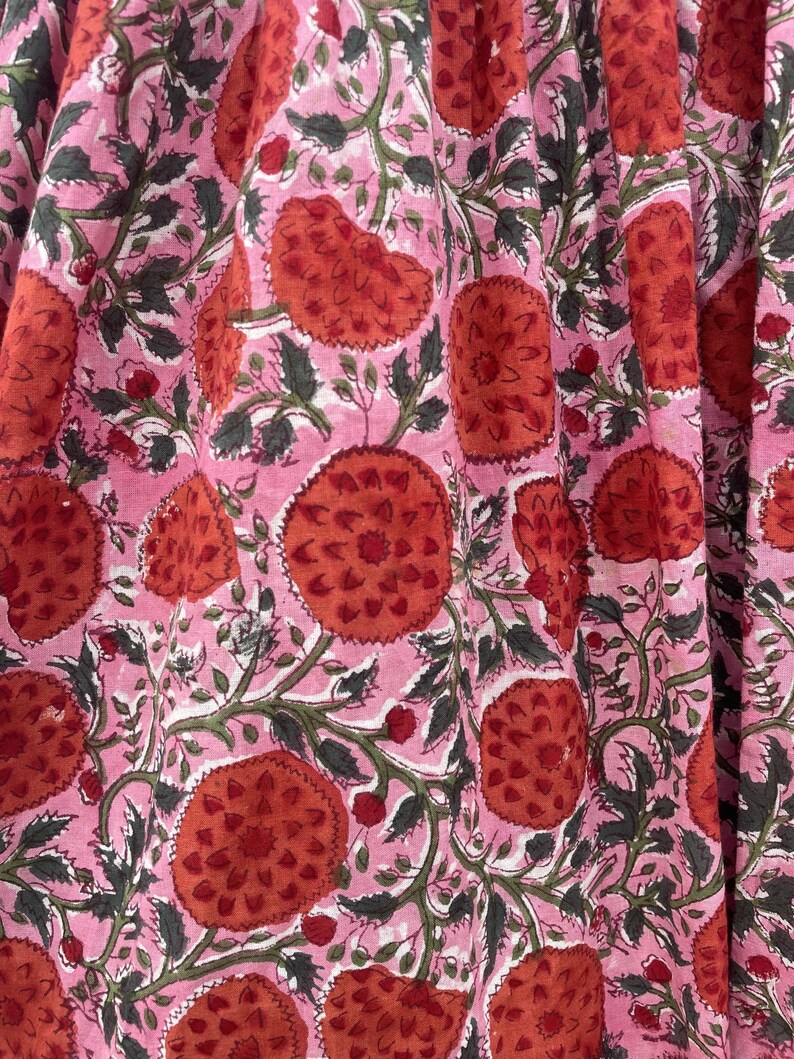 May include: A close-up of a pink fabric featuring a repeating pattern of large, stylized red flowers. The flowers have a textured appearance and are surrounded by green leaves and vines. The background is a soft pink.