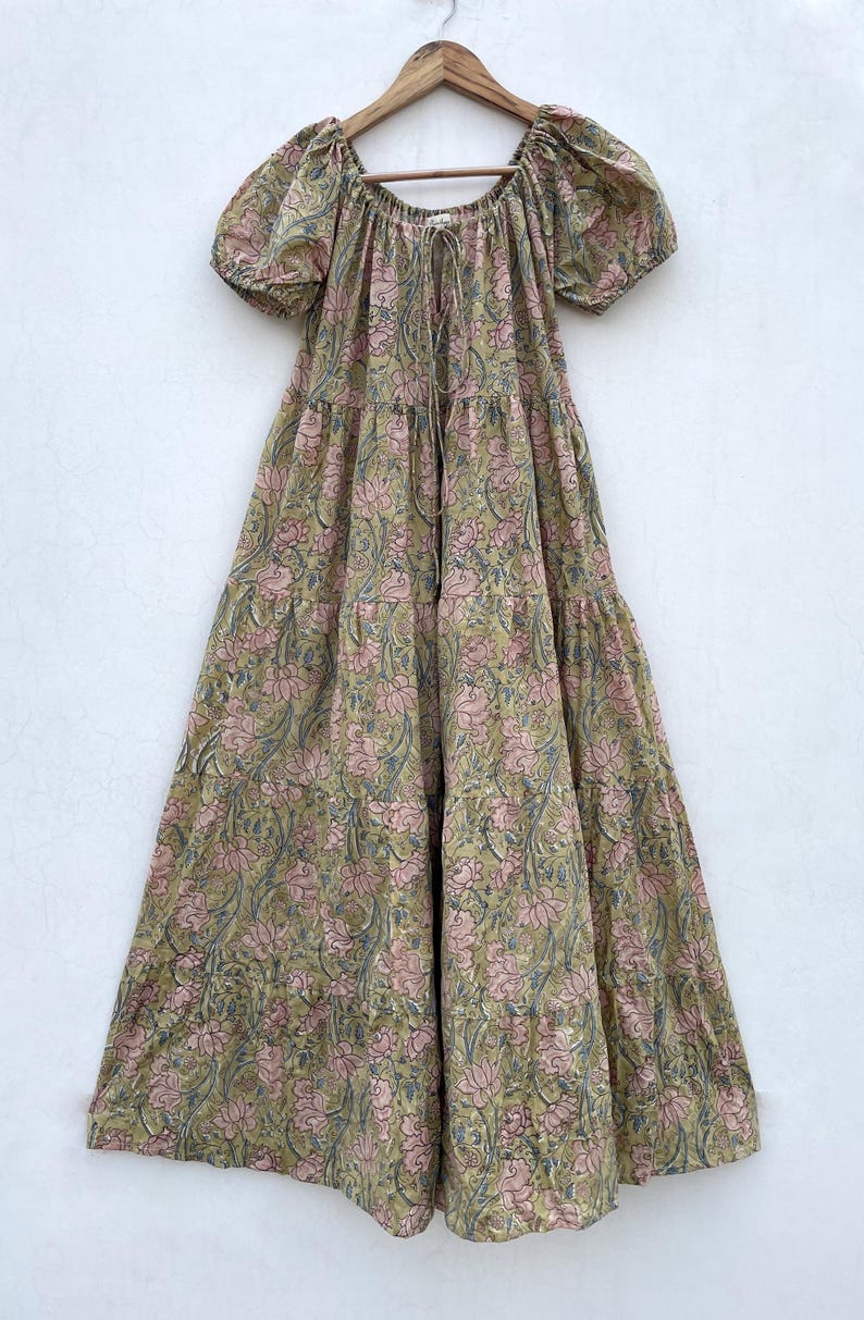 May include: A long, flowing dress with a floral pattern in shades of pink, green, and blue. The dress has short, puffed sleeves and a drawstring neckline. It is hanging on a wooden hanger.