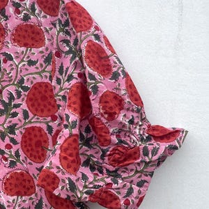 May include: Pink fabric with a repeating floral pattern in red and green. The fabric is gathered at the sleeve cuff, creating a ruffled edge. The floral design features large, stylized flowers and leaves.
