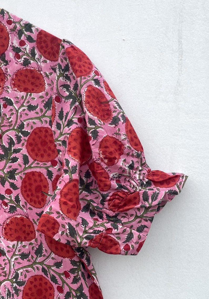 May include: Pink fabric with a repeating floral pattern in red and green. The fabric is gathered at the sleeve cuff, creating a ruffled edge. The floral design features large, stylized flowers and leaves.