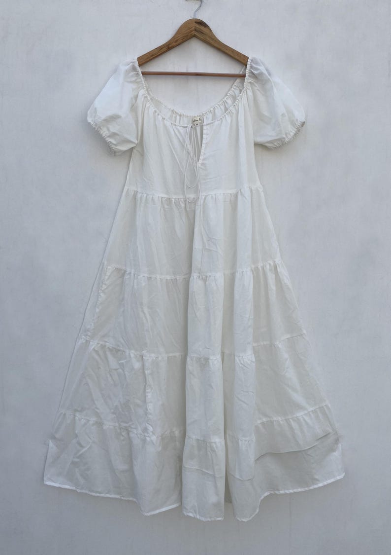 May include: A white tiered dress with short, puffed sleeves and a square neckline. The dress hangs on a wooden hanger against a plain white background. A drawstring detail is visible at the neckline.
