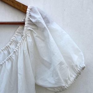 May include: A white, off-the-shoulder top with puffed sleeves, hanging on a wooden hanger. The fabric appears lightweight and slightly sheer, with gathered detailing along the neckline and sleeve cuffs. The top is likely made of cotton or a similar breathable material.