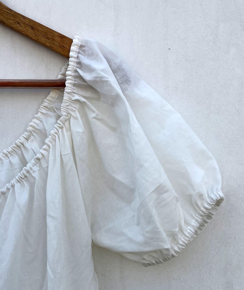 May include: A white, off-the-shoulder top with puffed sleeves, hanging on a wooden hanger. The fabric appears lightweight and slightly sheer, with gathered detailing along the neckline and sleeve cuffs. The top is likely made of cotton or a similar breathable material.