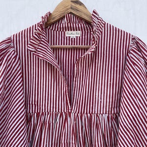 May include: A red and white striped cotton shirt with a ruffled collar and a V-neck. The shirt has a gathered waist and long sleeves. The label reads "Parichhan Fab" and "M".