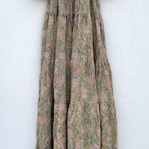 May include: A long, tiered dress with a floral pattern in shades of olive green, pink, and blue. The dress has short, puffed sleeves and a drawstring neckline. It hangs from a wooden hanger against a white background.
