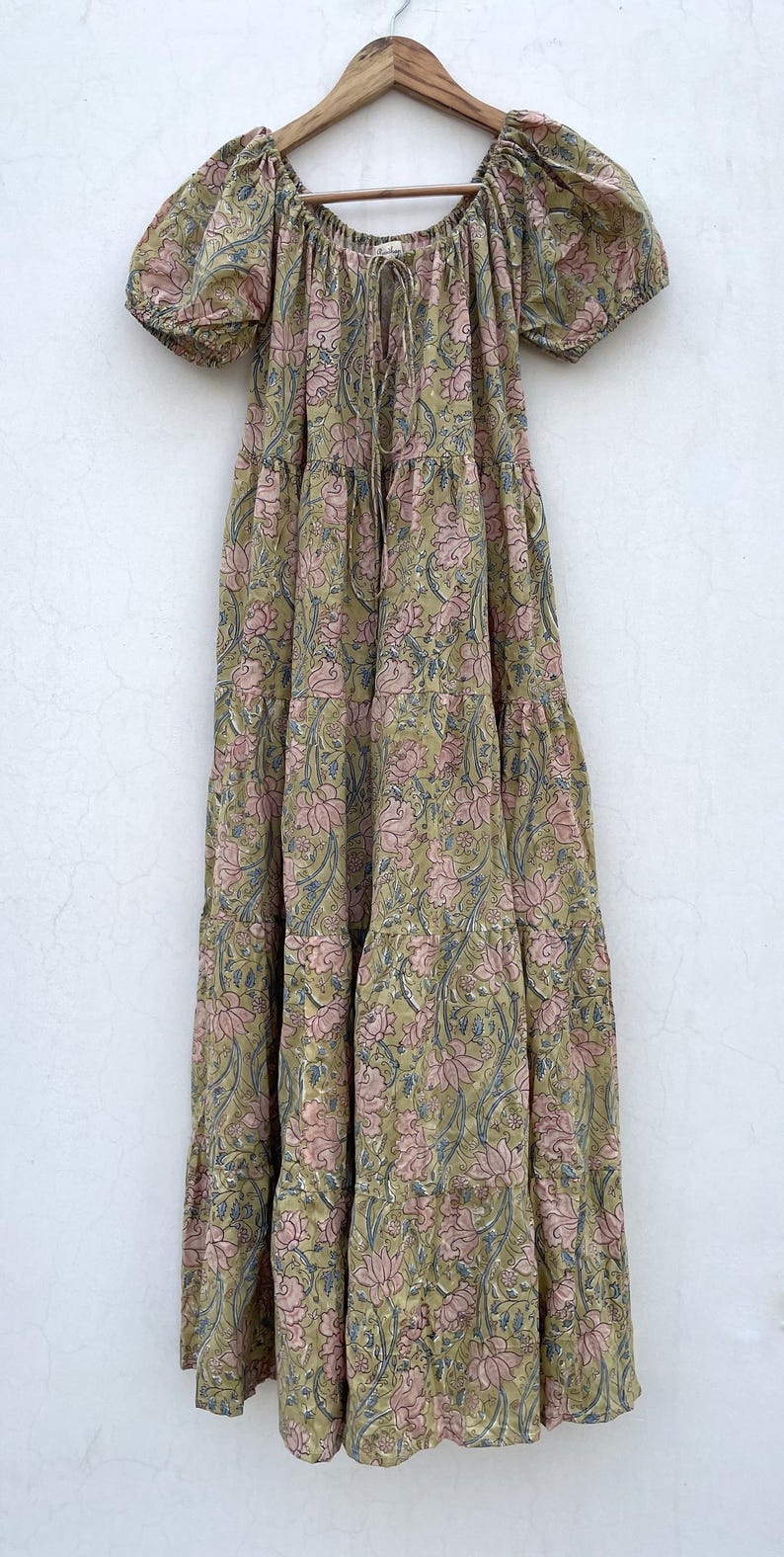 May include: A long, tiered dress with a floral pattern in shades of olive green, pink, and blue. The dress has short, puffed sleeves and a drawstring neckline. It hangs from a wooden hanger against a white background.