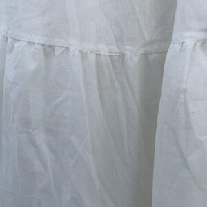 May include: Close-up of a white fabric garment with a tiered, ruffled design. The fabric appears to be lightweight and slightly textured, with visible seams and folds. The overall aesthetic is soft and flowing.
