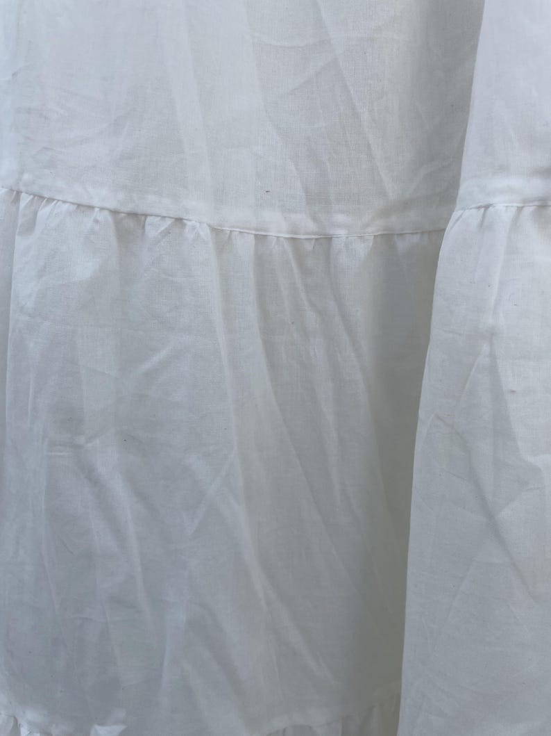 May include: Close-up of a white fabric garment with a tiered, ruffled design. The fabric appears to be lightweight and slightly textured, with visible seams and folds. The overall aesthetic is soft and flowing.