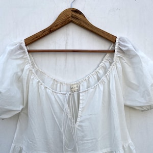 May include: A white, off-the-shoulder top with short, puffy sleeves, hanging on a wooden hanger. The top has a square neckline and a drawstring detail at the chest. The fabric appears to be lightweight cotton.