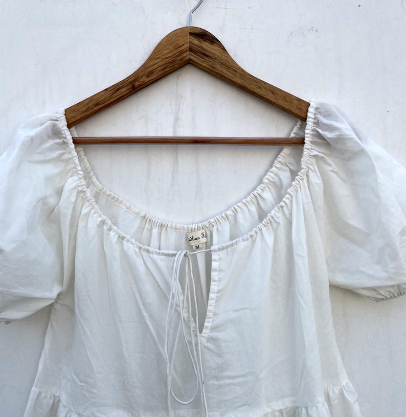 May include: A white, off-the-shoulder top with short, puffy sleeves, hanging on a wooden hanger. The top has a square neckline and a drawstring detail at the chest. The fabric appears to be lightweight cotton.