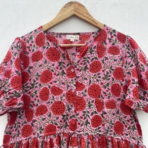 May include: A pink dress with a red fruit pattern and green leaves, hanging on a wooden hanger. The dress features short sleeves, a button-down front, and a tiered skirt. The fabric has a floral design.