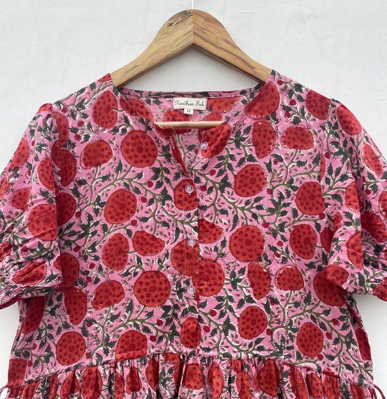 May include: A pink dress with a red fruit pattern and green leaves, hanging on a wooden hanger. The dress features short sleeves, a button-down front, and a tiered skirt. The fabric has a floral design.
