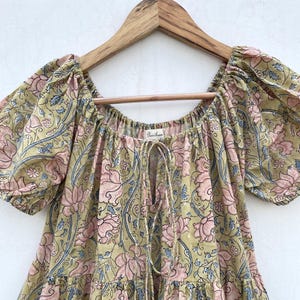 May include: A floral print dress in shades of olive green, pink, and blue, hanging on a wooden hanger. The dress features a square neckline, short puff sleeves, and a drawstring detail. The fabric is patterned with large floral motifs.