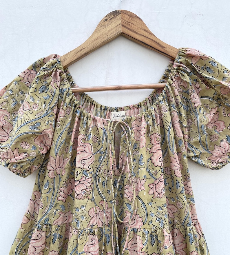 May include: A floral print dress in shades of olive green, pink, and blue, hanging on a wooden hanger. The dress features a square neckline, short puff sleeves, and a drawstring detail. The fabric is patterned with large floral motifs.