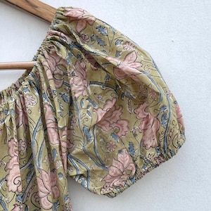 May include: Close-up of a floral patterned sleeve. The fabric features a repeating design of pink flowers and blue leaves on a light olive green background. The sleeve is gathered at the cuff and shoulder.