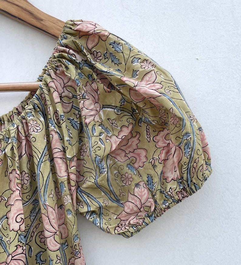May include: Close-up of a floral patterned sleeve. The fabric features a repeating design of pink flowers and blue leaves on a light olive green background. The sleeve is gathered at the cuff and shoulder.