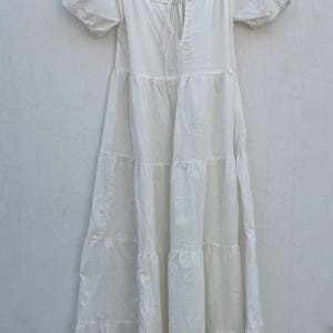May include: A white tiered dress with short, puffed sleeves hangs from a wooden hanger. The dress features a square neckline with a drawstring detail and a flowing, tiered skirt. The fabric appears lightweight and suitable for warm weather.