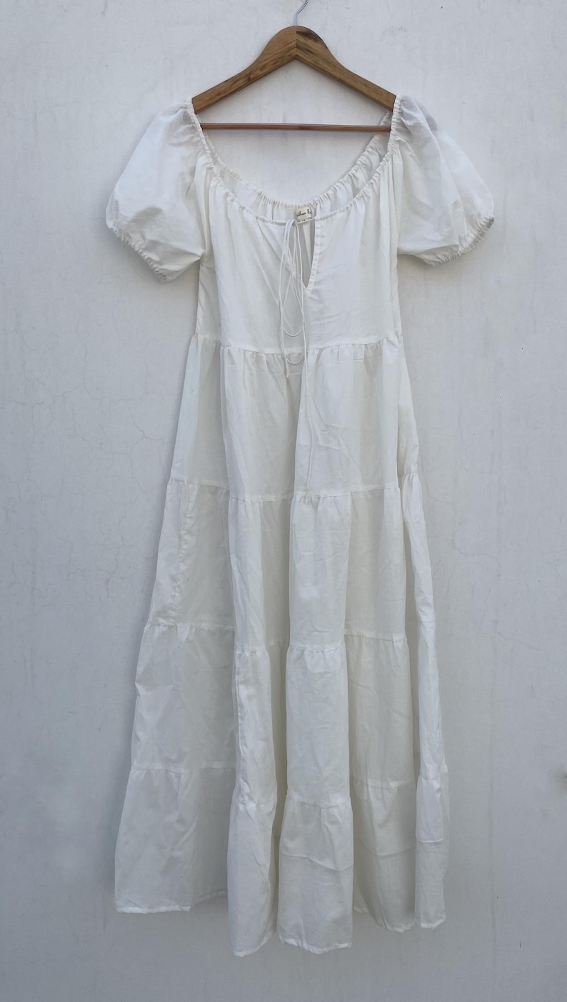 May include: A white tiered dress with short, puffed sleeves hangs from a wooden hanger. The dress features a square neckline with a drawstring detail and a flowing, tiered skirt. The fabric appears lightweight and suitable for warm weather.