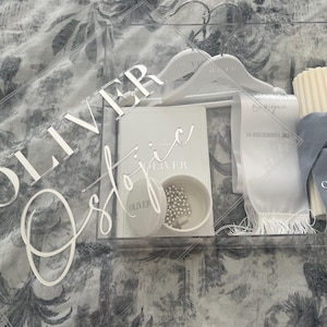 May include: A clear acrylic box contains white hangers, a small book, a bowl with silver beads, a candle, and a blue ribbon. The box has the name "Oliver" in large, white, stylized letters. The items are arranged on a patterned surface.