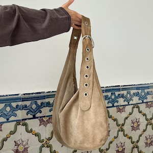 Op de afbeelding: Een beige su&egrave;de hobo-tas met een enkele schouderriem. De riem is aan de tas bevestigd met een grote zilveren metalen ring en grommets. De tas heeft een zachte, slordige vorm.