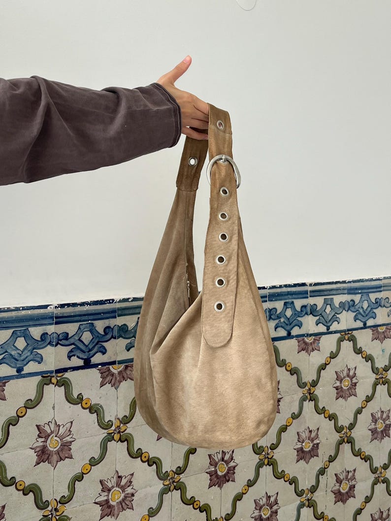 Op de afbeelding: Een beige su&egrave;de hobo-tas met een enkele schouderriem. De riem is aan de tas bevestigd met een grote zilveren metalen ring en grommets. De tas heeft een zachte, slordige vorm.