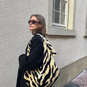 Op de afbeelding: Een zwarte jas met een grote schoudertas met een cr&egrave;me- en zwart zebraprint. De tas heeft een zachte, pluche textuur. De persoon draagt een donkere zonnebril en oorringen. De achtergrond is een lichtgekleurde, getextureerde muur.