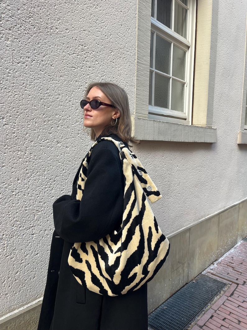 Op de afbeelding: Een zwarte jas met een grote schoudertas met een cr&egrave;me- en zwart zebraprint. De tas heeft een zachte, pluche textuur. De persoon draagt een donkere zonnebril en oorringen. De achtergrond is een lichtgekleurde, getextureerde muur.