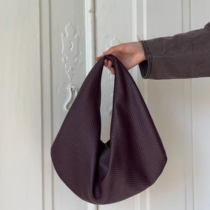 Op de afbeelding: Een diepe bordeauxrode handtas met een gestructureerd, gewatteerd patroon. De tas heeft een enkele handgreep en een zachte, losse vorm. De achtergrond is een witte deur.