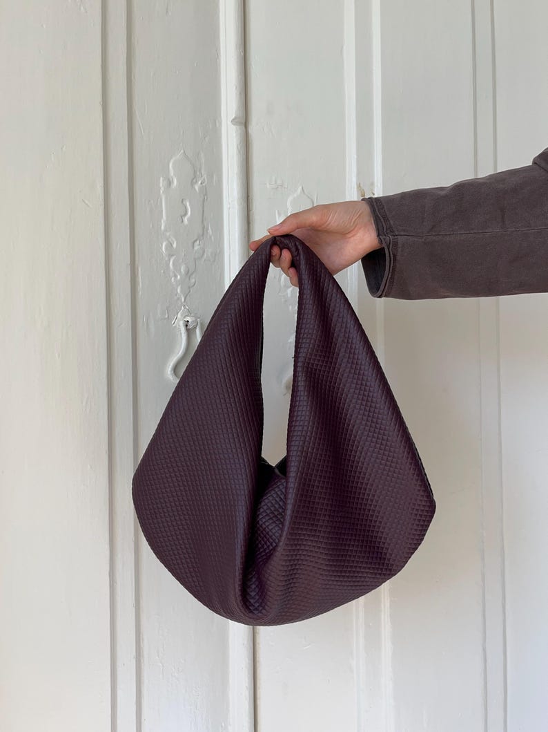 Op de afbeelding: Een diepe bordeauxrode handtas met een gestructureerd, gewatteerd patroon. De tas heeft een enkele handgreep en een zachte, losse vorm. De achtergrond is een witte deur.