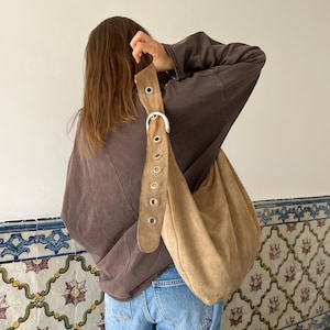 Op de afbeelding: Een beige su&egrave;de schoudertas met een grote gesp en riemdetails. De tas wordt gedragen over een bruin sweatshirt en een blauwe spijkerbroek. De tas heeft een nonchalant ontwerp.