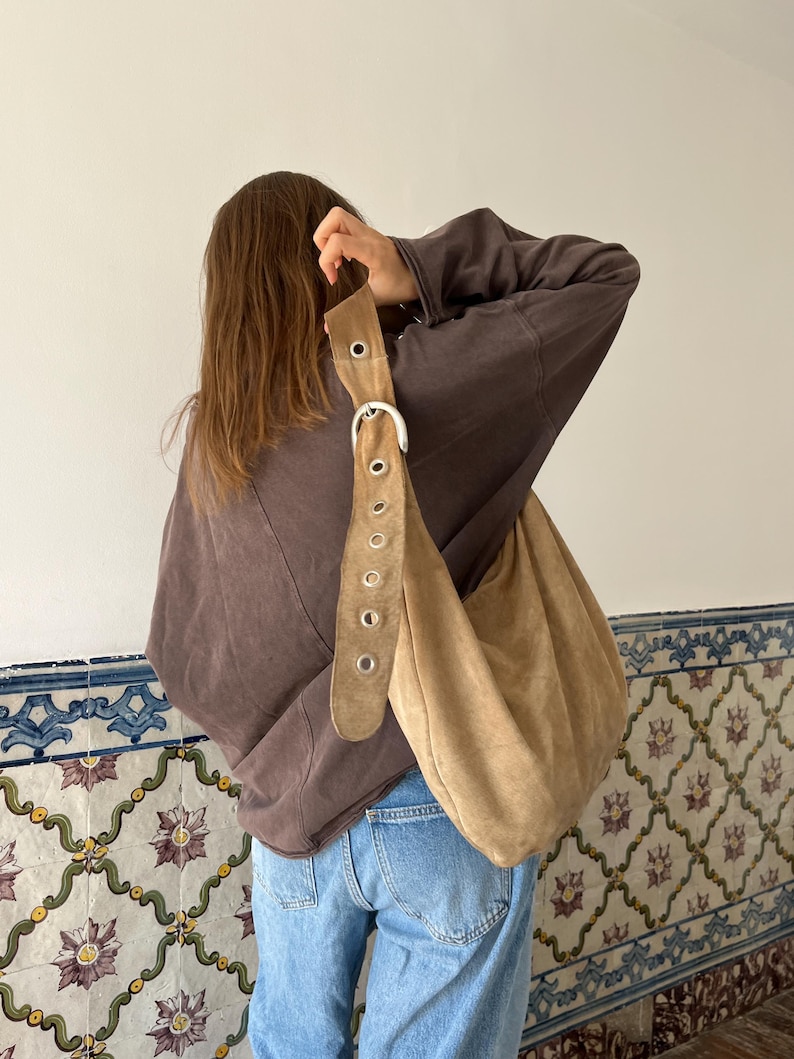 Op de afbeelding: Een beige su&egrave;de schoudertas met een grote gesp en riemdetails. De tas wordt gedragen over een bruin sweatshirt en een blauwe spijkerbroek. De tas heeft een nonchalant ontwerp.