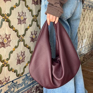 Op de afbeelding: Een bordeauxrode gewatteerde handtas met een unieke, zachte vorm. De tas wordt vastgehouden door een persoon die een blauwe spijkerbroek en een bruine sweatshirt draagt. De achtergrond heeft een decoratieve tegelwand met bloemmotieven.