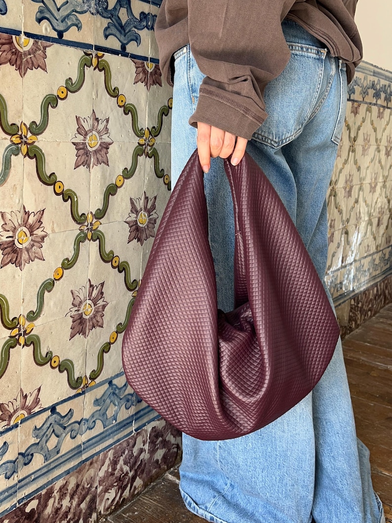 Op de afbeelding: Een bordeauxrode gewatteerde handtas met een unieke, zachte vorm. De tas wordt vastgehouden door een persoon die een blauwe spijkerbroek en een bruine sweatshirt draagt. De achtergrond heeft een decoratieve tegelwand met bloemmotieven.