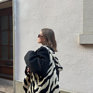 Op de afbeelding: Een zwart-witte zebraprint tote bag met een bijpassende capuchon. De tas wordt over de schouder gedragen. De persoon draagt een zwarte jas en een zonnebril. De achtergrond is een witte muur met textuur.