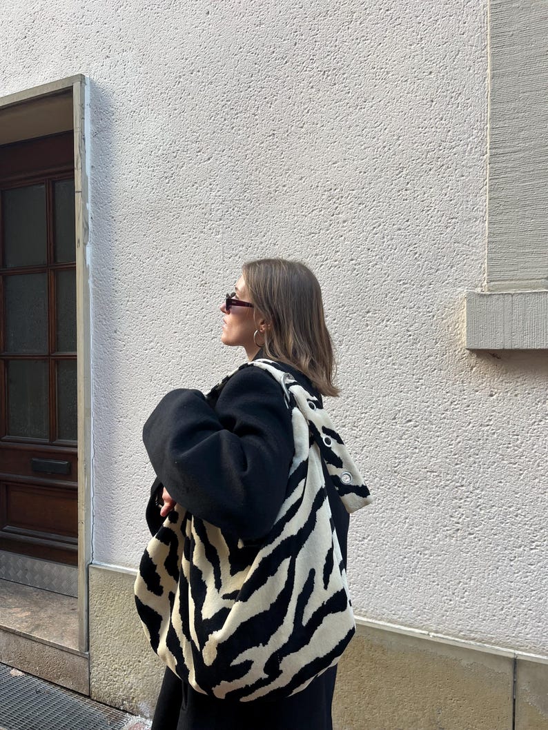 Op de afbeelding: Een zwart-witte zebraprint tote bag met een bijpassende capuchon. De tas wordt over de schouder gedragen. De persoon draagt een zwarte jas en een zonnebril. De achtergrond is een witte muur met textuur.