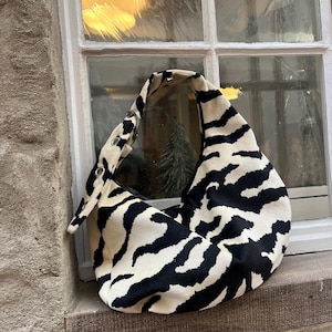 Op de afbeelding: Een schoudertas met zwart-witte zebraprint en een gebogen ontwerp. De tas is gemaakt van een zacht materiaal en heeft een enkele band met metalen oogjes. De tas ligt op een stenen vensterbank.