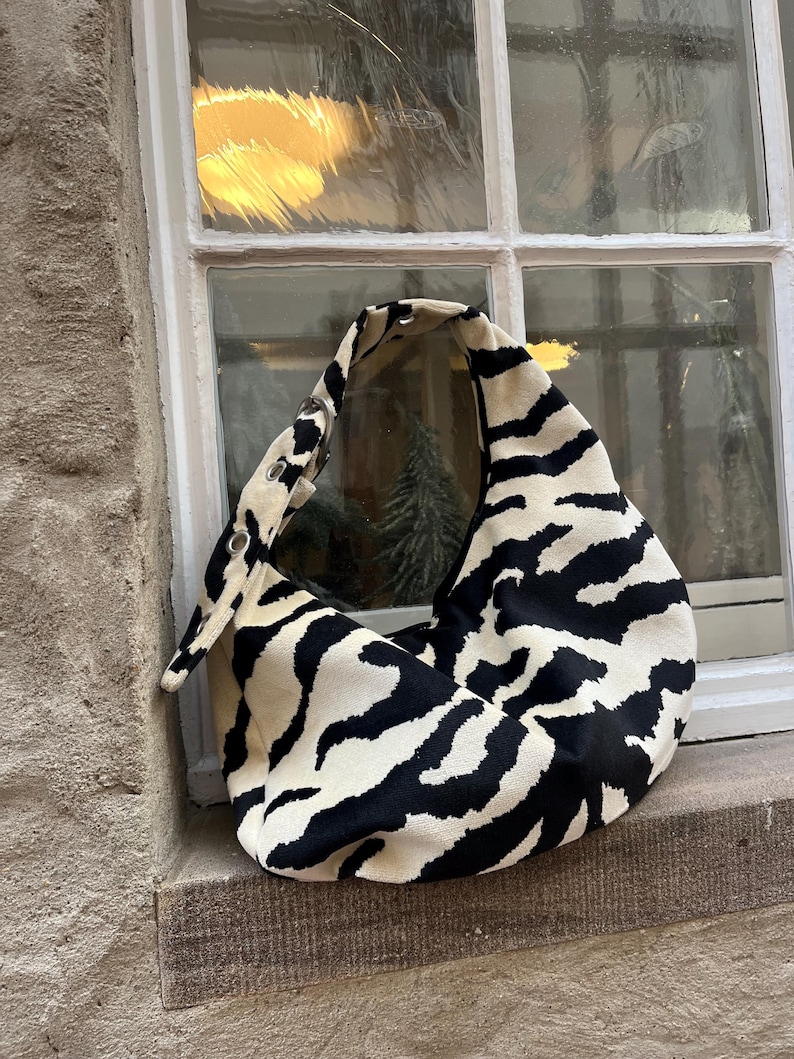 Op de afbeelding: Een schoudertas met zwart-witte zebraprint en een gebogen ontwerp. De tas is gemaakt van een zacht materiaal en heeft een enkele band met metalen oogjes. De tas ligt op een stenen vensterbank.