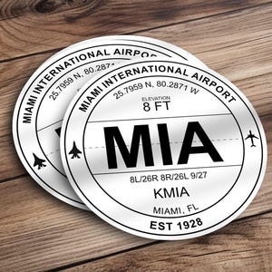May include: White round sticker with black text that reads "Miami International Airport" and "MIA". The sticker also includes the airport code "KMIA", the city "Miami, FL", and the year "EST 1928".