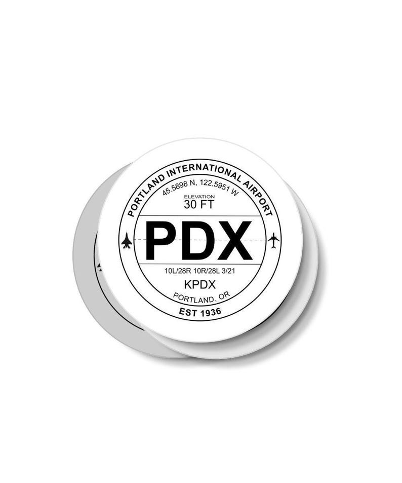 May include: A white round sticker with black text and a black airplane symbol. The sticker says "Portland International Airport", "45.5898 N. 122.5951 W", "Elevation 30 FT", "PDX", "10L/28R 10R/28L 3/21", "KPDX", "Portland, OR", and "Est 1936".