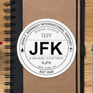 May include: A white round sticker with black text that reads "John F. Kennedy International Airport" with the airport code "JFK" and the airport's elevation of 13 feet. The sticker also includes the airport's runway numbers and the city and state "New York, NY".
