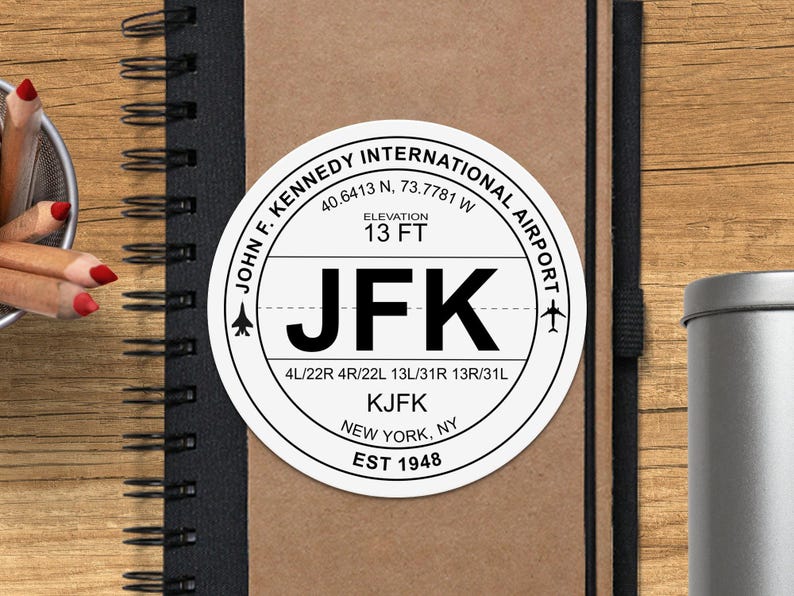 May include: A white round sticker with black text that reads "John F. Kennedy International Airport" with the airport code "JFK" and the airport's elevation of 13 feet. The sticker also includes the airport's runway numbers and the city and state "New York, NY".