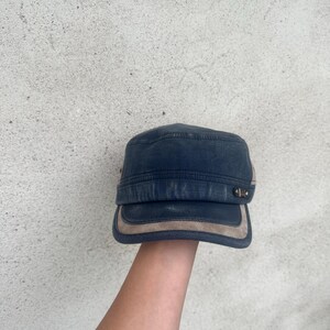 May include: A navy blue military-style cap with a tan suede-like brim and accents. The cap has a flat top and a short brim. The hat is made of a durable fabric and features metal hardware on the side.
