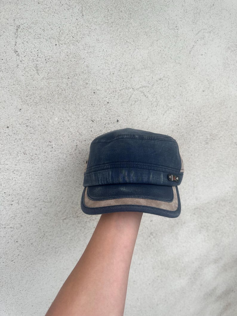 May include: A navy blue military-style cap with a tan suede-like brim and accents. The cap has a flat top and a short brim. The hat is made of a durable fabric and features metal hardware on the side.